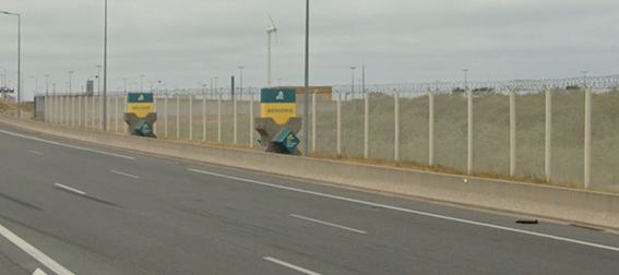 A road into Calais with tall fences topped with razor wire and barbed wire. In front of the fences are signs that read 'Welcome' and 'Bienvenue'.